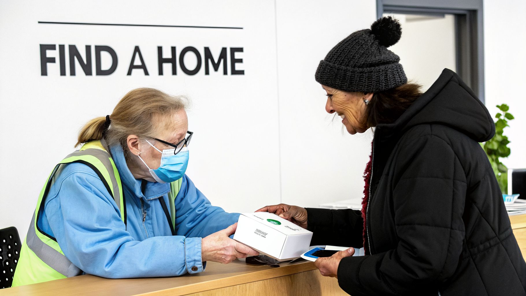 A masked volunteer hands a white box to a smiling woman in a black coat and beanie.